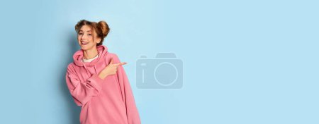 Téléchargez les photos : Jeune fille positive à capuche rose posant, pointant avec excitation sur fond bleu studio. Bonne nouvelle. Flyer. Concept de jeunesse, beauté, mode, mode de vie, émotions, expression faciale. Publicité - en image libre de droit