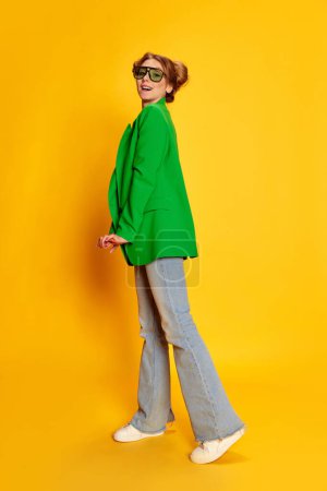 Téléchargez les photos : Portrait de jeune fille en veste vert vif posant sur fond jaune studio. Une jeunesse élégante et à la mode. Concept de jeunesse, beauté, mode, mode de vie, émotions, expression faciale. Publicité - en image libre de droit