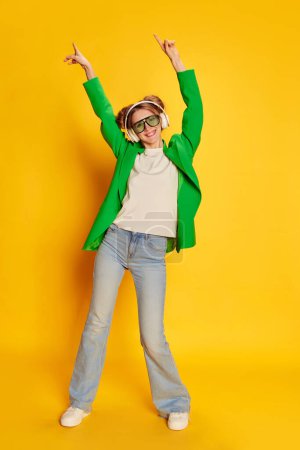 Foto de Retrato de niña en chaqueta verde brillante posando en auriculares sobre fondo de estudio amarillo. Emocionado. Concepto de juventud, belleza, moda, estilo de vida, emociones, expresión facial. Anuncio - Imagen libre de derechos