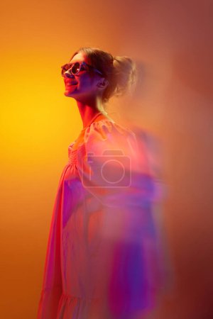 Photo for Portrait of young beautiful girl in white dress posing with smile over orange background in neon lights. Unfocused effect. Concept of youth, beauty, fashion, lifestyle, emotions, facial expression. Ad - Royalty Free Image