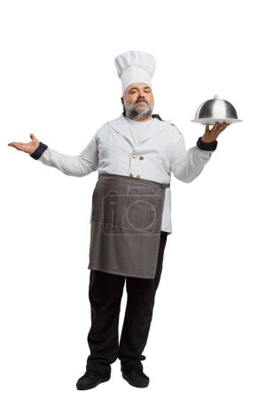 Téléchargez les photos : Portrait d'homme barbu, chef de restaurant en uniforme servant un plat isolé sur fond blanc. Elle a l'air fière. Concept de profession, profession, passe-temps, mode de vie, goût. Publicité - en image libre de droit