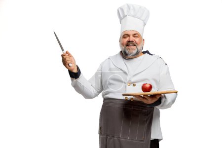 Foto de Retrato de hombre barbudo alegre, chef de restaurante en uniforme con cuchillo y tomate sobre tabla de cortar aislado sobre fondo blanco. Concepto de profesión, ocupación, afición, estilo de vida, gusto. Anuncio - Imagen libre de derechos
