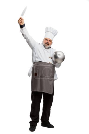 Téléchargez les photos : Portrait d'homme barbu, chef de restaurant en uniforme posant avec couteau et bol isolé sur fond blanc. Prêt à travailler. Concept de profession, profession, passe-temps, mode de vie, goût. Publicité - en image libre de droit