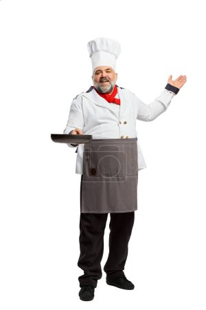 Téléchargez les photos : Portrait d'homme barbu, chef de restaurant en uniforme posant avec poêle isolée sur fond blanc. Cuisiner. Concept de profession, profession, passe-temps, mode de vie, goût. Publicité - en image libre de droit