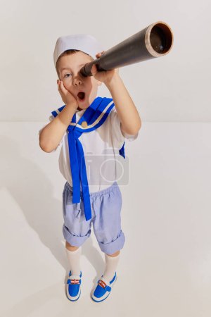 Téléchargez les photos : Portrait de petit garçon, enfant à l'image d'un marin en gilet posant avec du verre espion sur fond gris. Impressionné. Concept d'enfance, amitié, plaisir, mode de vie, mode, voyage, émotions - en image libre de droit