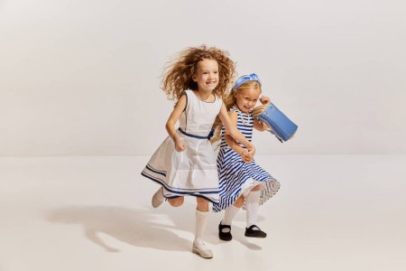 Foto de Retrato de dos niñas, niños con vestidos lindos corriendo alegremente, posando sobre fondo gris. Feliz. Concepto de infancia, amistad, diversión, estilo de vida, moda, estilo retro, emociones - Imagen libre de derechos