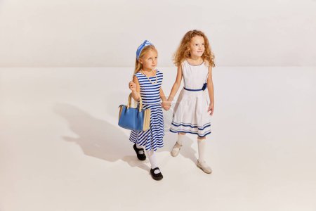 Photo for Portrait of two little girls, children in cute dresses holding hands and posing over grey background. Cruise style. Concept of childhood, friendship, fun, lifestyle, fashion, retro style, emotions - Royalty Free Image