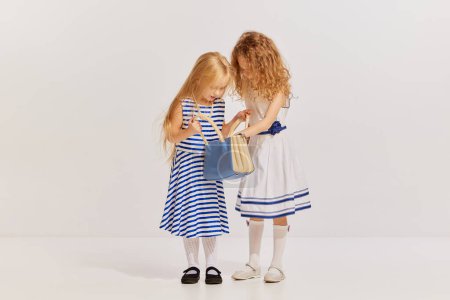 Téléchargez les photos : Portrait de deux petites filles, enfants en robes mignonnes posant sur fond gris. Regarde à l'intérieur du sac. Concept d'enfance, amitié, plaisir, style de vie, mode, style rétro, émotions - en image libre de droit