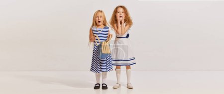 Téléchargez les photos : Portrait de deux petites filles, enfants en robes mignonnes posant avec des visages choqués sur fond gris. Surprise. Concept d'enfance, amitié, plaisir, style de vie, mode, style rétro, émotions - en image libre de droit