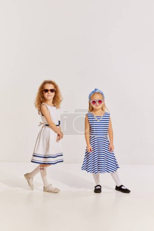 Téléchargez les photos : Portrait de deux petites filles, des enfants en robes mignonnes et des lunettes de soleil à la mode posant sur fond gris. Voyageurs. Concept d'enfance, amitié, plaisir, style de vie, mode, style rétro, émotions - en image libre de droit