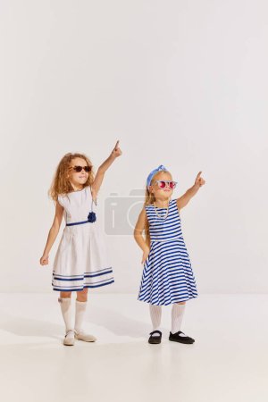 Téléchargez les photos : Portrait de deux petites filles, des enfants en robes mignonnes et des lunettes de soleil posant, pointant sur fond gris. Voyage. Concept d'enfance, amitié, plaisir, style de vie, mode, style rétro, émotions - en image libre de droit