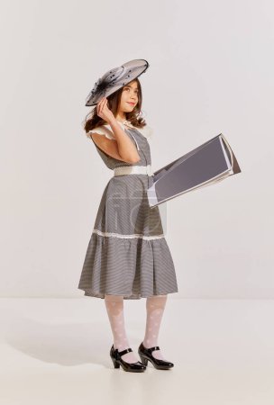 Photo for Portrait of stylish teen girl in vintage dress and hat posing with magazine over grey background. 50s fashion look. Concept of childhood, friendship, fun, lifestyle, fashion, retro style, emotions - Royalty Free Image