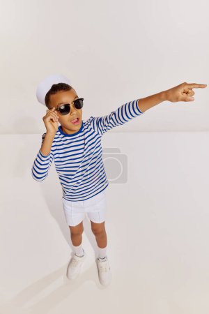 Téléchargez les photos : Portrait de garçon, enfant en gilet rayé et lunettes posant, pointant sur fond gris. Vacances de croisière. Concept d'enfance, amitié, plaisir, style de vie, mode, style rétro, émotions - en image libre de droit