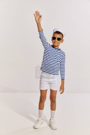 Téléchargez les photos : Portrait de garçon joyeux, enfant en gilet rayé et lunettes de soleil posant sur fond gris. Vacances d'été. Concept d'enfance, amitié, plaisir, style de vie, mode, style rétro, émotions - en image libre de droit