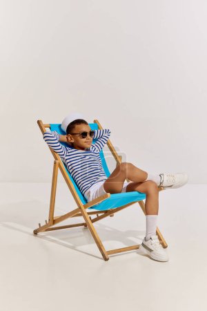 Foto de Retrato de niño, niño en chaleco a rayas y gafas de sol sentado en la silla, posando sobre fondo gris. Viajero de mar. Concepto de infancia, amistad, diversión, estilo de vida, moda, estilo retro, emociones - Imagen libre de derechos