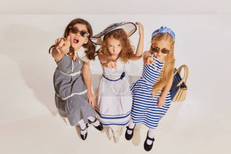 Téléchargez les photos : Trois belles filles, des enfants assis, posant dans des robes élégantes et des lunettes de soleil sur fond gris. Concept d'enfance, amitié, plaisir, style de vie, mode, style rétro, émotions - en image libre de droit