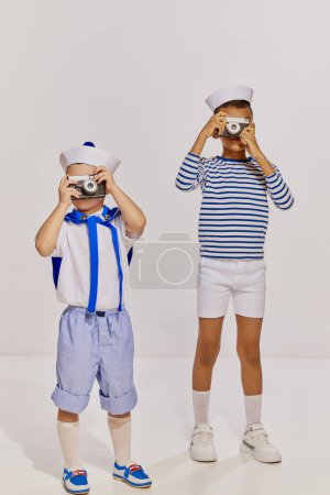 Téléchargez les photos : Portrait de deux garçons, des enfants dans des cotumes de style mer, gilet photo avec des appareils photo vintage sur fond gris. Concept d'enfance, amitié, plaisir, style de vie, mode, style rétro, émotions - en image libre de droit