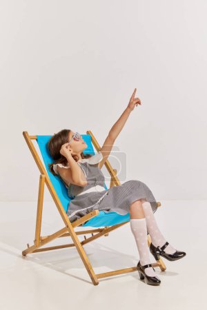 Téléchargez les photos : Portrait de belle adolescente en robe vintage posant, assise sur une chaise et pointant sur un fond gris. Croisière. Concept d'enfance, amitié, plaisir, style de vie, mode, style rétro, émotions - en image libre de droit