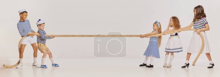 Téléchargez les photos : Portrait d'enfants posant dans des vêtements de style marin, tirant la corde, jouant sur fond gris. Concept d'enfance, amitié, plaisir, style de vie, mode, style rétro, émotions - en image libre de droit