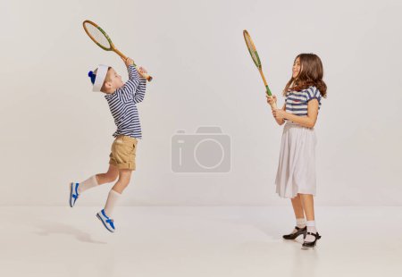 Téléchargez les photos : Portrait d'enfants, garçon et fille dans des vêtements de style marin jouant au badminton, posant sur fond gris. Concept d'enfance, amitié, plaisir, style de vie, mode, style rétro, émotions - en image libre de droit
