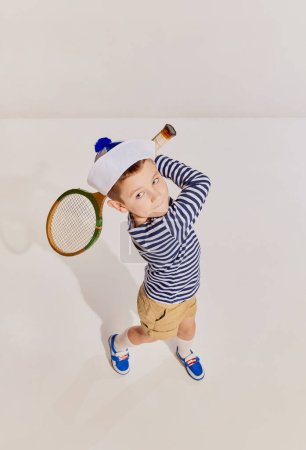 Foto de Retrato de niño, niño en chaleco a rayas posando con raqueta vintage, jugando bádminton sobre fondo gris. Concepto de infancia, amistad, diversión, estilo de vida, moda, estilo retro, emociones - Imagen libre de derechos