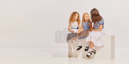 Téléchargez les photos : Trois belles filles, des enfants assis, posant dans des robes mignonnes, lisant magazine sur fond gris. Concept d'enfance, amitié, plaisir, style de vie, mode, style rétro, émotions - en image libre de droit