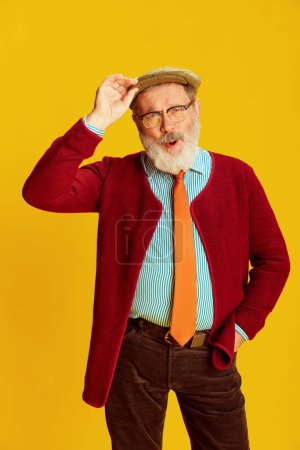Téléchargez les photos : Portrait de l'homme âgé émotif en vêtements classiques, lunettes et casquette posant sur un fond jaune vif. Ambiance positive. Concept d'émotions, expression faciale, style de vie, mode moderne - en image libre de droit