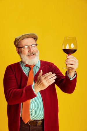 Téléchargez les photos : Portrait de l'homme âgé en vêtements classiques, verres et casquette posant, verre montant avec du vin rouge sur fond jaune vif. Sommelier. Concept d'émotions, expression faciale, style de vie, mode moderne - en image libre de droit