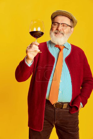 Foto de Retrato del hombre mayor en ropa clásica, vasos y gorra posando con copa de vino tinto sobre fondo amarillo vivo. Amante del gusto. Concepto de emociones, expresión facial, estilo de vida, moda moderna - Imagen libre de derechos