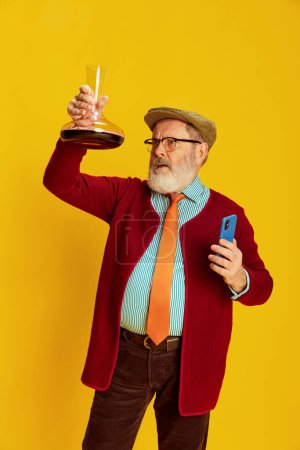 Foto de Retrato de hombre mayor en ropa clásica, vasos y posando gorra, mirando n decantador de vino sobre fondo amarillo vivo. Experto en vino. Concepto de emociones, expresión facial, estilo de vida, moda moderna - Imagen libre de derechos