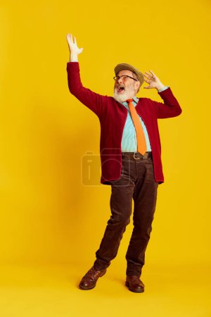 Foto de Retrato de un hombre mayor con ropa clásica, gafas y gorra posando sobre un fondo amarillo vivo. Emoción positiva. Concepto de emociones, expresión facial, estilo de vida, moda moderna - Imagen libre de derechos