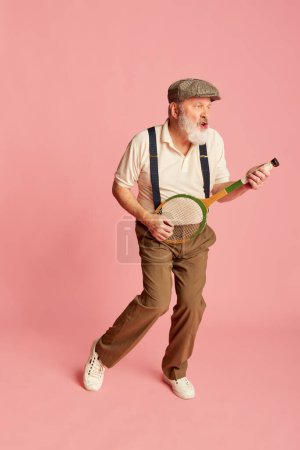 Téléchargez les photos : Portrait de vieil homme âgé dans des vêtements élégants jouant avec la raquette comme la guitare sur fond rose. Loisirs. Concept d'émotions, expression faciale, style de vie, mode moderne - en image libre de droit