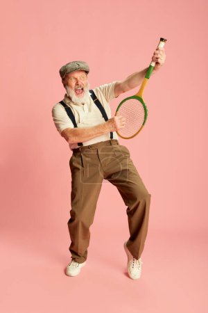 Foto de Retrato de anciano con ropa clásica elegante posando con raqueta, sobre fondo rosa. Ilusión de guitarra. Concepto de emociones, expresión facial, estilo de vida, moda moderna - Imagen libre de derechos