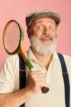 Téléchargez les photos : Portrait d'homme âgé drôle en vêtements classiques posant avec raquette de badminton vintage sur fond rose. Loisirs. Concept d'émotions, expression faciale, style de vie, mode moderne - en image libre de droit