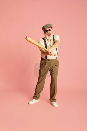 Photo for Portrait of stylish senior man in classical clothes, cap and sunglasses posing with bat over pink background. Serving. Baseball lover. Concept of emotions, facial expression, lifestyle, modern fashion - Royalty Free Image