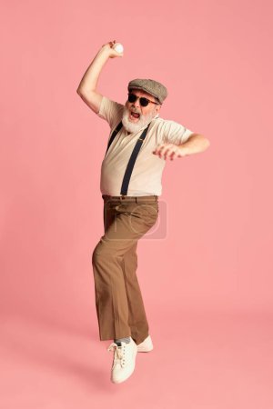 Photo for Portrait of stylish senior man in classical clothes posing, emotionally throwing baseball ball over pink background. Concept of emotions, facial expression, lifestyle, modern fashion - Royalty Free Image