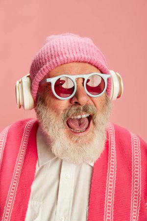 Téléchargez les photos : Portrait de sénior élégant, vieil homme en tenue lumineuse élégante avec chapeau tricoté et lunettes de soleil posant dans des écouteurs sur fond rose. Concept d'émotions, expression faciale, style de vie, mode moderne - en image libre de droit