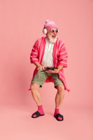 Téléchargez les photos : Portrait de sénior élégant, vieil homme dans une tenue élégante et lumineuse avec des lunettes de soleil et des écouteurs posant sur fond rose. Hipster. Concept d'émotions, expression faciale, style de vie, mode moderne - en image libre de droit