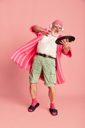 Téléchargez les photos : Portrait de sénior élégant, vieil homme dans une tenue lumineuse élégante avec du vinyle posant sur fond rose. Club, dj. Concept d'émotions, expression faciale, style de vie, mode moderne - en image libre de droit