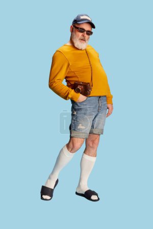 Téléchargez les photos : Portrait d'un homme élégant, âgé et barbu en vêtements décontractés avec casquette, lunettes de soleil et caméra vintage posant sur fond bleu. Concept d'émotions, expression faciale, style de vie, mode moderne - en image libre de droit