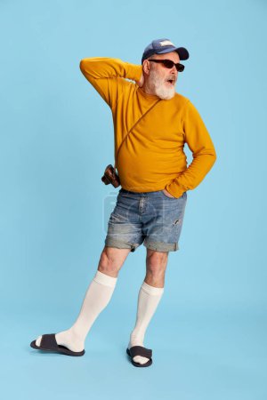 Téléchargez les photos : Portrait d'homme barbu senior en vêtements décontractés avec casquette, lunettes de soleil et caméra vintage posant sur fond bleu. Loisirs. Concept d'émotions, expression faciale, style de vie, mode moderne - en image libre de droit
