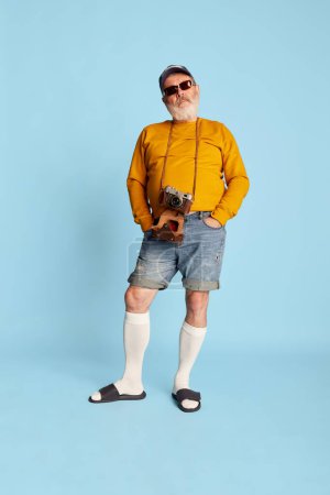 Téléchargez les photos : Portrait d'homme barbu senior en vêtements décontractés avec casquette, lunettes de soleil et caméra vintage posant sur fond bleu. Voyage. Concept d'émotions, expression faciale, style de vie, mode moderne - en image libre de droit