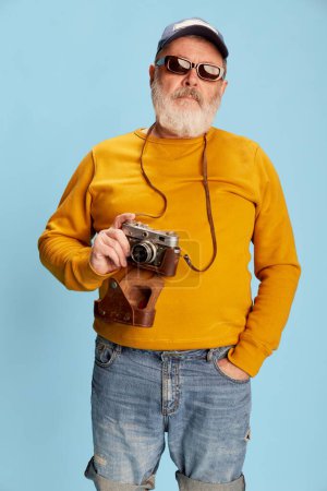 Téléchargez les photos : Portrait d'homme barbu senior en vêtements décontractés avec casquette, lunettes de soleil et caméra vintage posant sur fond bleu. Touristique. Concept d'émotions, expression faciale, style de vie, mode moderne - en image libre de droit