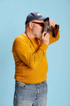 Photo for Senior bearded man in casual clothes with cap, sunglasses, taking photo with vintage camera posing over blue background. Tourist. Concept of emotions, facial expression, lifestyle, modern fashion - Royalty Free Image
