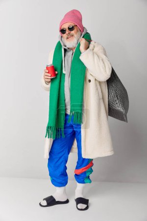Foto de Retrato de anciano con estilo, hombre viejo con abrigo de piel, sombrero rosa de punto y bufanda verde posando sobre fondo gris. Concepto de emociones, expresión facial, estilo de vida, moda moderna - Imagen libre de derechos