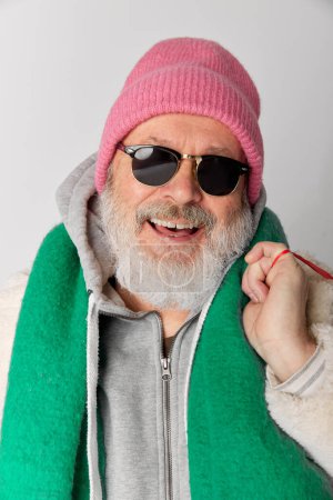 Foto de Retrato de hombre mayor con estilo, anciano de punto sombrero rosa y bufanda verde posando en gafas de sol sobre fondo gris. El invierno. Concepto de emociones, expresión facial, estilo de vida, moda moderna - Imagen libre de derechos