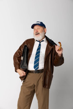 Téléchargez les photos : Portrait de vieil homme âgé en veste en cuir posant avec mallette et parapluie sur fond gris. Elle a l'air joyeuse. Concept d'émotions, expression faciale, style de vie, mode moderne - en image libre de droit