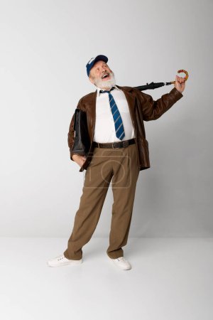 Téléchargez les photos : Portrait de vieil homme âgé en veste en cuir posant avec mallette et parapluie sur fond gris. Joyeux rire. Concept d'émotions, expression faciale, style de vie, mode moderne - en image libre de droit