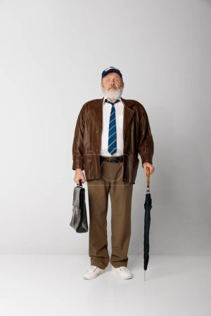 Téléchargez les photos : Portrait de vieil homme âgé en veste en cuir posant avec mallette et parapluie sur fond gris. Employé. Je regarde droit. Concept d'émotions, expression faciale, style de vie, mode moderne - en image libre de droit