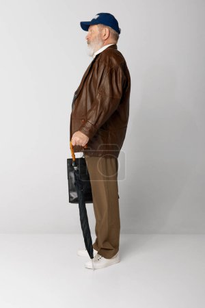 Téléchargez les photos : Portrait de vieil homme âgé en veste en cuir posant avec mallette et parapluie sur fond gris. Employé. Vue latérale. Concept d'émotions, expression faciale, style de vie, mode moderne - en image libre de droit